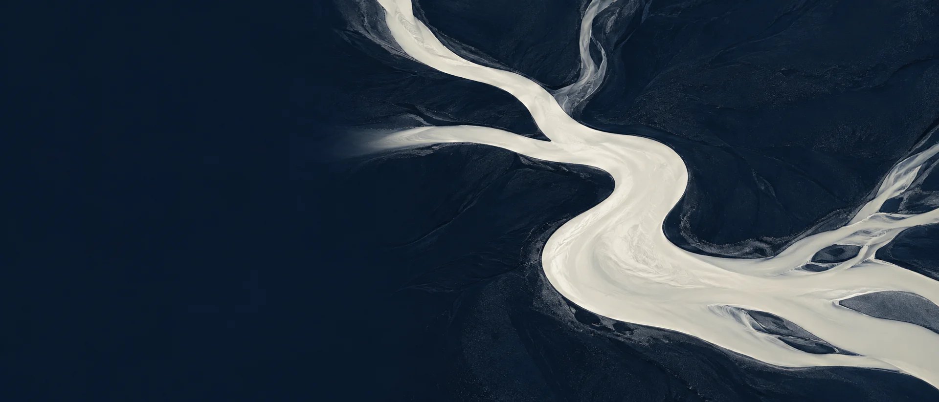 Aerial photograph of a braided glacial river: cream silt water winding in an S-bend across dark volcanic ground